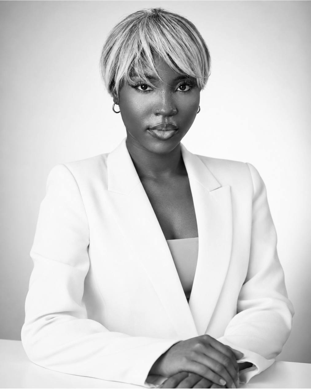 Divine in a white blazer, seated at a desk with her hands folded
